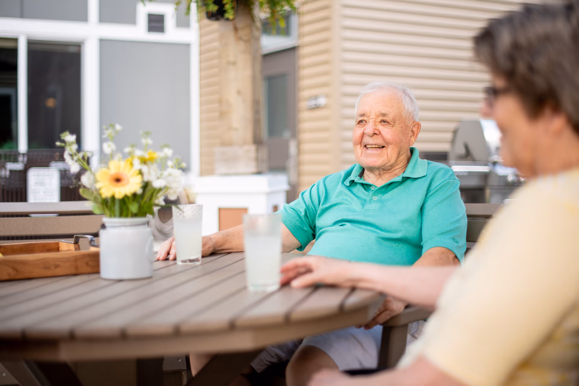 An elderly man in a green polo shirt sitting at an outdoor table with a woman, both enjoying a drink. There is a vase with flowers on the table and the setting appears to be a patio area of a senior living facility.