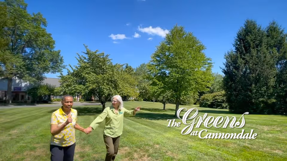Two people holding hands and walking across a sunny grassy lawn with trees and a building in the background and the words "The Greens at Cannondale" overlaid.
