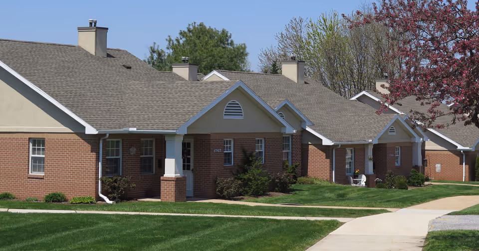 Row of single-story brick residential buildings with beige roofs and white trim, surrounded by green lawns and trees with some blooming flowers under a clear blue sky.