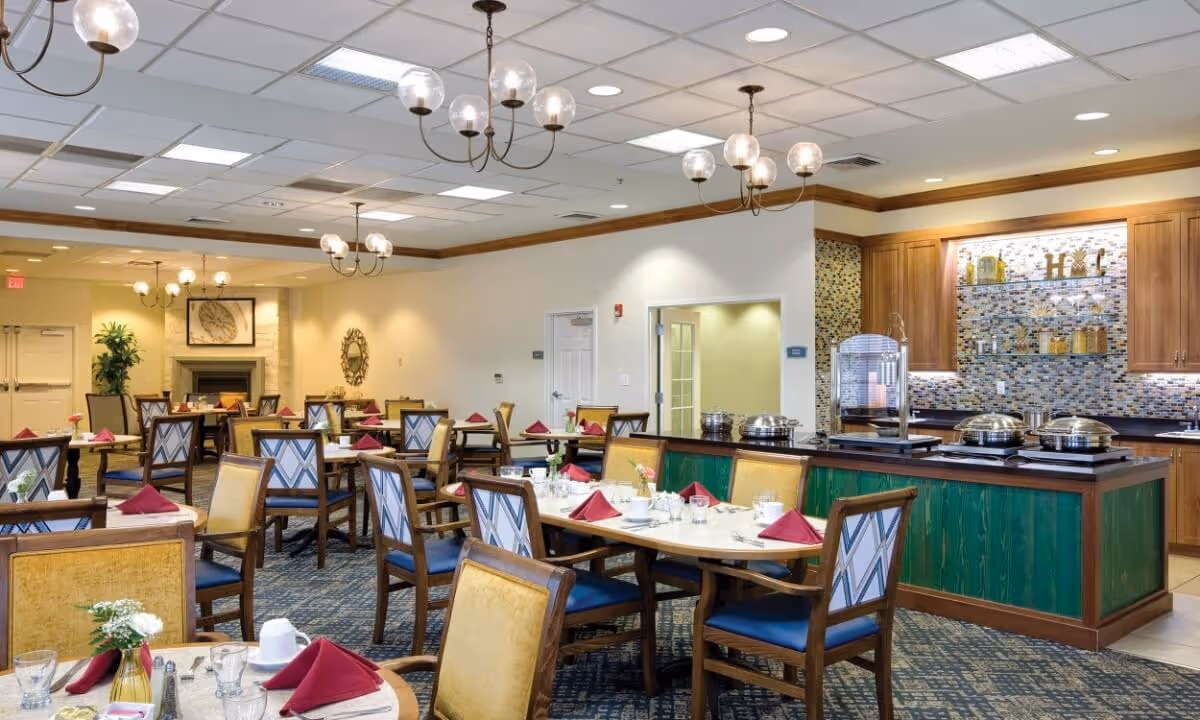 A spacious dining room with multiple tables set with white tablecloths, red folded napkins, glassware, and silverware. The room features wooden chairs with blue and yellow upholstery, chandeliers with globe lights hanging from the ceiling, and a buffet area with covered serving dishes and a decorative tiled backsplash.