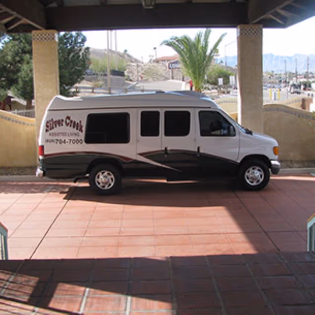 A white and black shuttle van parked under a covered area with tiled flooring. The van has the text 'Silver Creek Assisted Living' and a phone number on its side. In the background, there are palm trees, a beige wall, and a mountainous landscape.