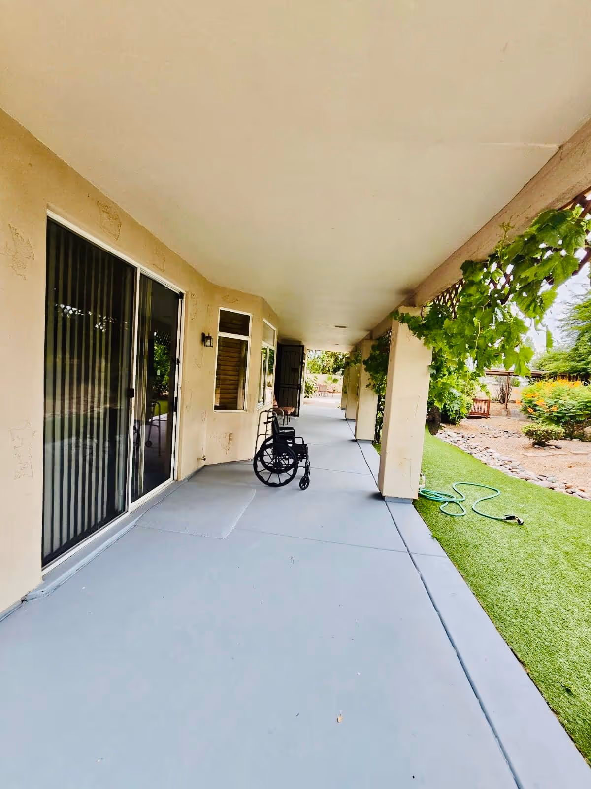 Covered outdoor walkway with beige walls and ceiling, a wheelchair positioned near a sliding glass door, green plants hanging from the ceiling beams, and a garden hose on artificial grass next to a landscaped area with bushes and rocks.