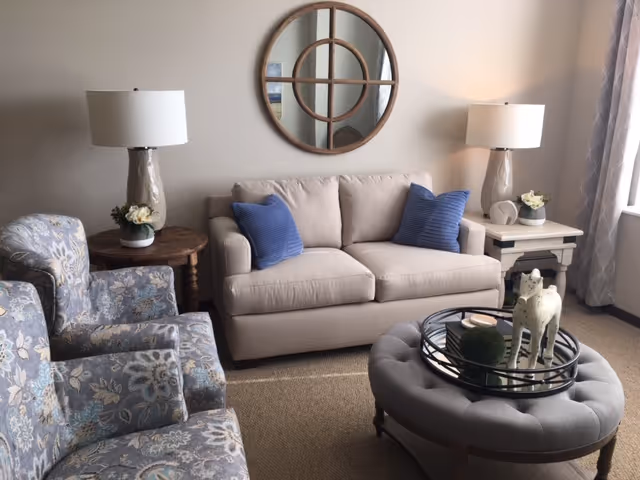 Cozy living room with a beige loveseat flanked by side tables and lamps, two patterned armchairs, a round wall mirror, and a tufted ottoman with a decorative tray.