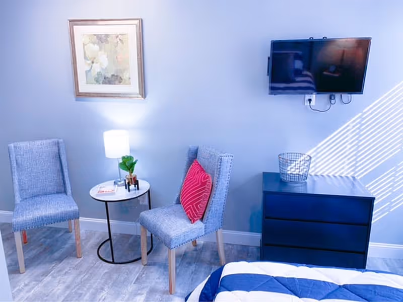 A cozy bedroom corner with two gray upholstered chairs, one with a red decorative pillow, a small round side table with a lamp and a small plant, a framed floral painting on the wall, a mounted flat-screen TV, and a dark blue dresser with a wire basket on top. Part of a bed with a blue and white comforter is visible in the foreground.