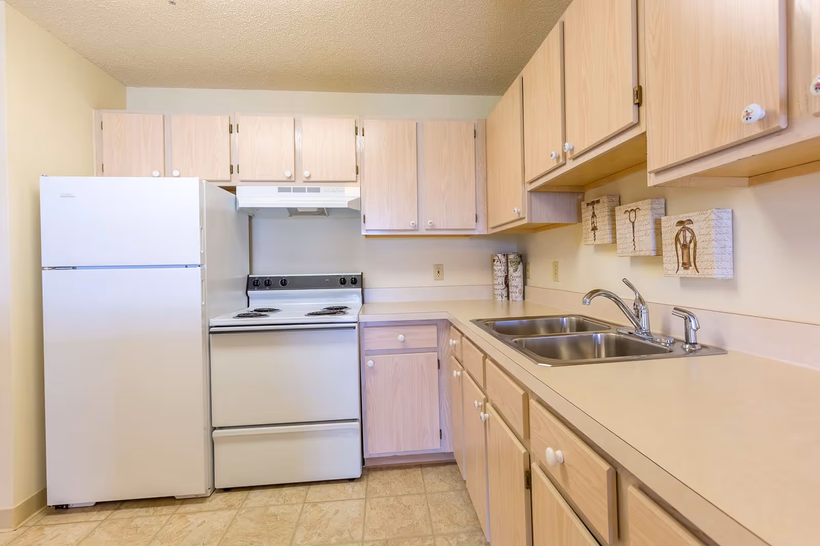 A compact kitchen with light wood cabinets, a white refrigerator, an electric stove with a range hood, a double stainless steel sink, and beige countertops. The walls are light-colored, and there are three decorative wall hangings above the sink area.