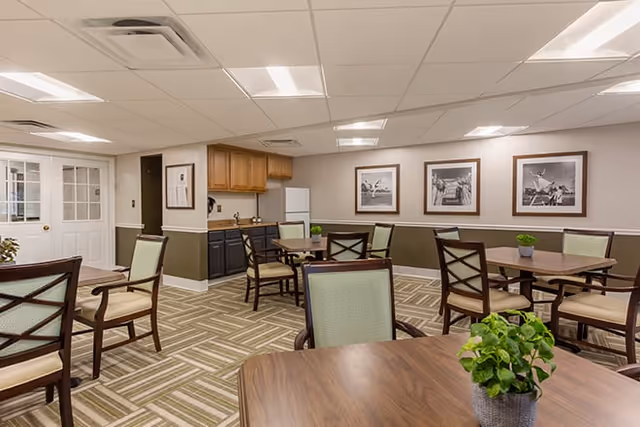 A bright communal dining area with several wooden tables and upholstered chairs, potted plants, framed photos on the wall, and a small kitchenette.