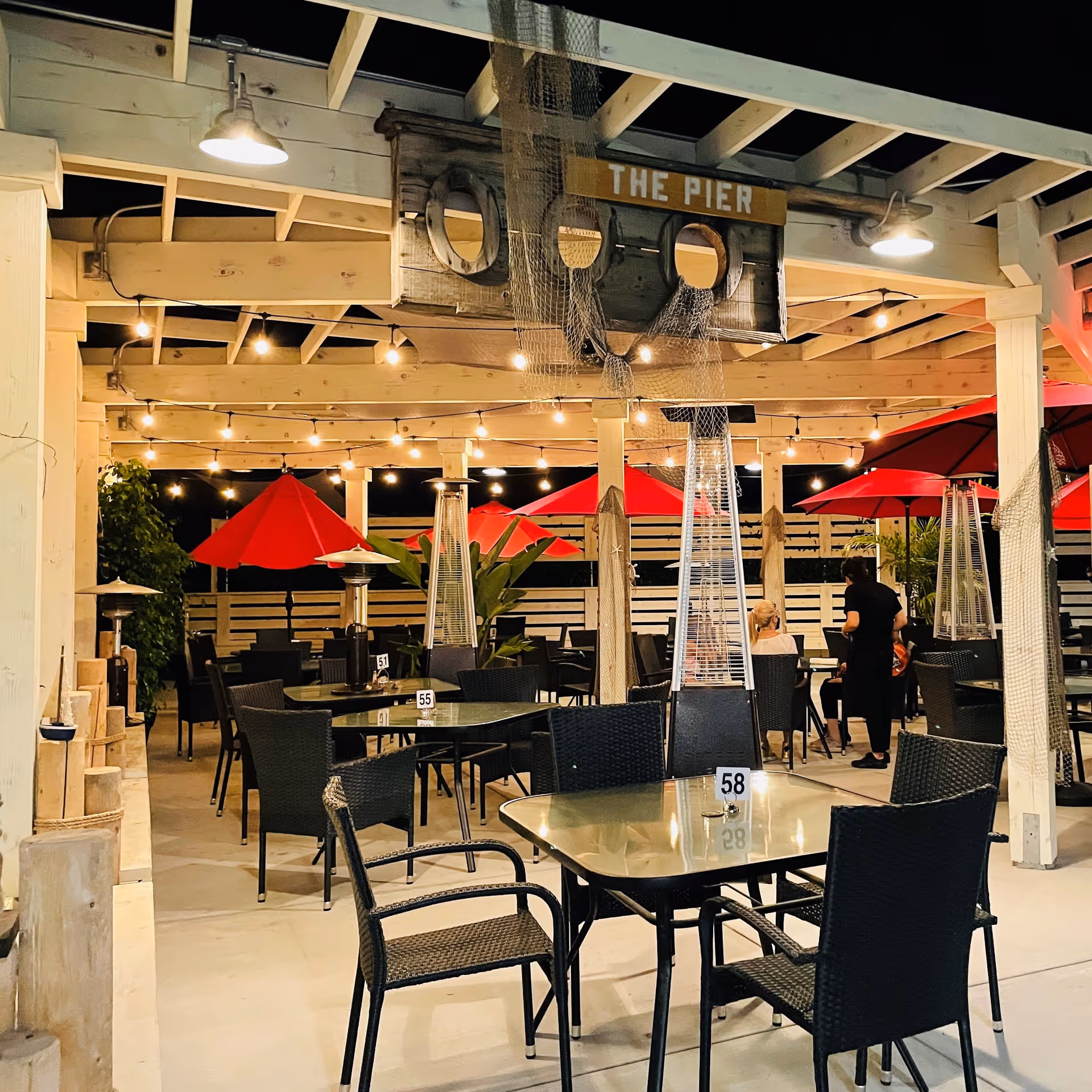 Outdoor dining area at night with tables and chairs under a wooden pergola. Red umbrellas and string lights create a cozy atmosphere. A wooden sign reading 'THE PIER' hangs from the pergola, and there are tall outdoor heaters near the tables. Some people are seated and a server is attending to them.