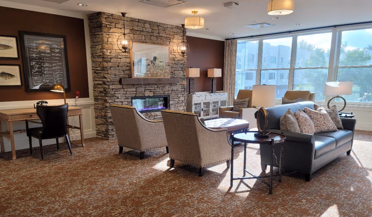 A cozy common area in a senior living facility featuring comfortable seating, a stone fireplace, and large windows.