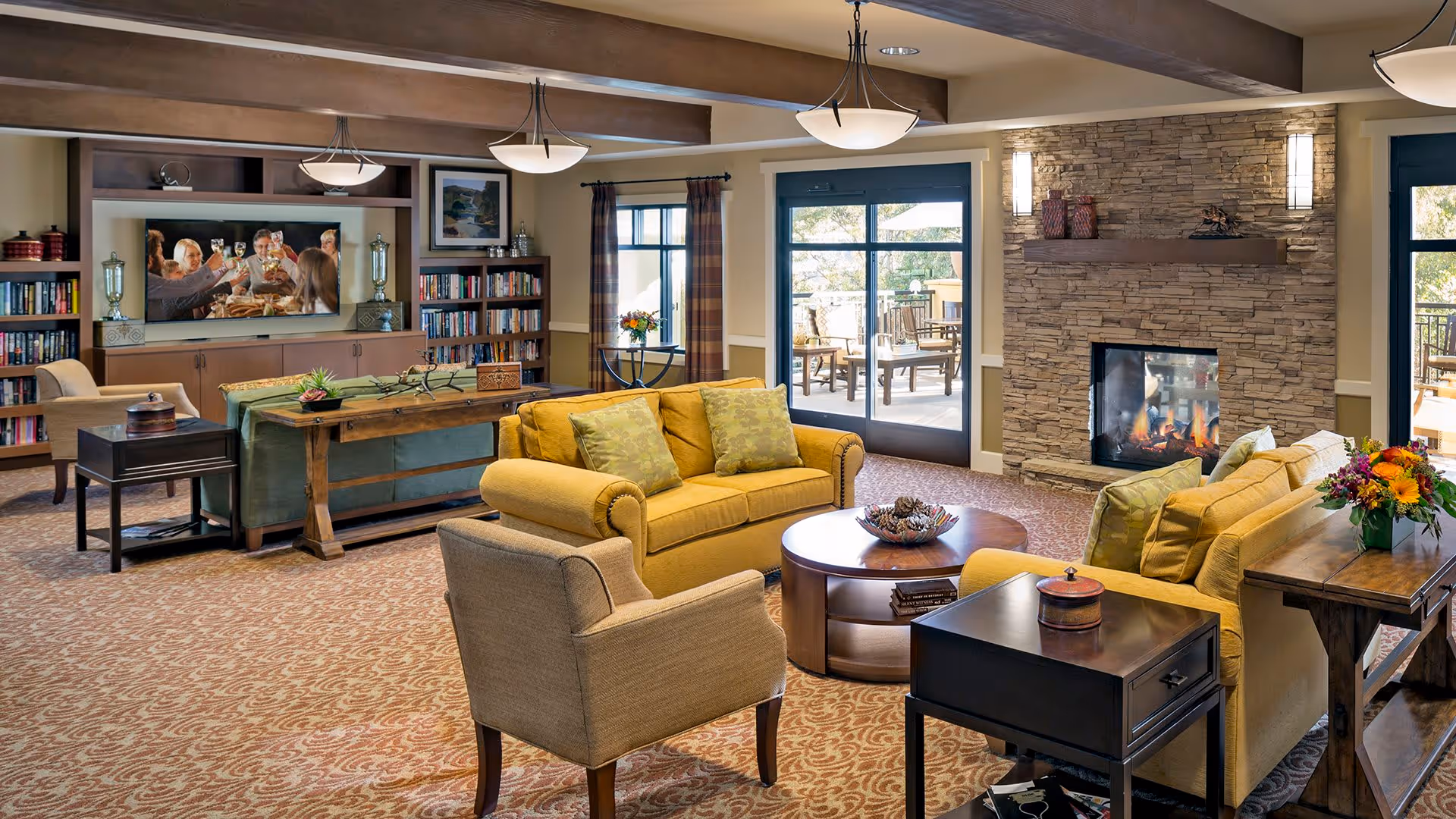 A cozy living room in a senior living facility with yellow sofas, beige armchairs, a round wooden coffee table, and a stone fireplace with a fire burning. The room has carpeted flooring, bookshelves, a large TV showing people toasting, and large windows and glass doors leading to an outdoor patio with seating.