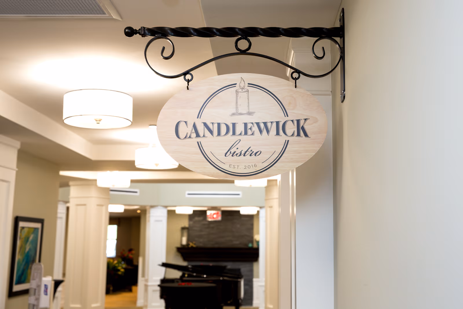 A wooden hanging sign with the text 'CANDLEWICK bistro EST. 2016' displayed in an interior hallway with white walls, ceiling lights, and a piano in the background.