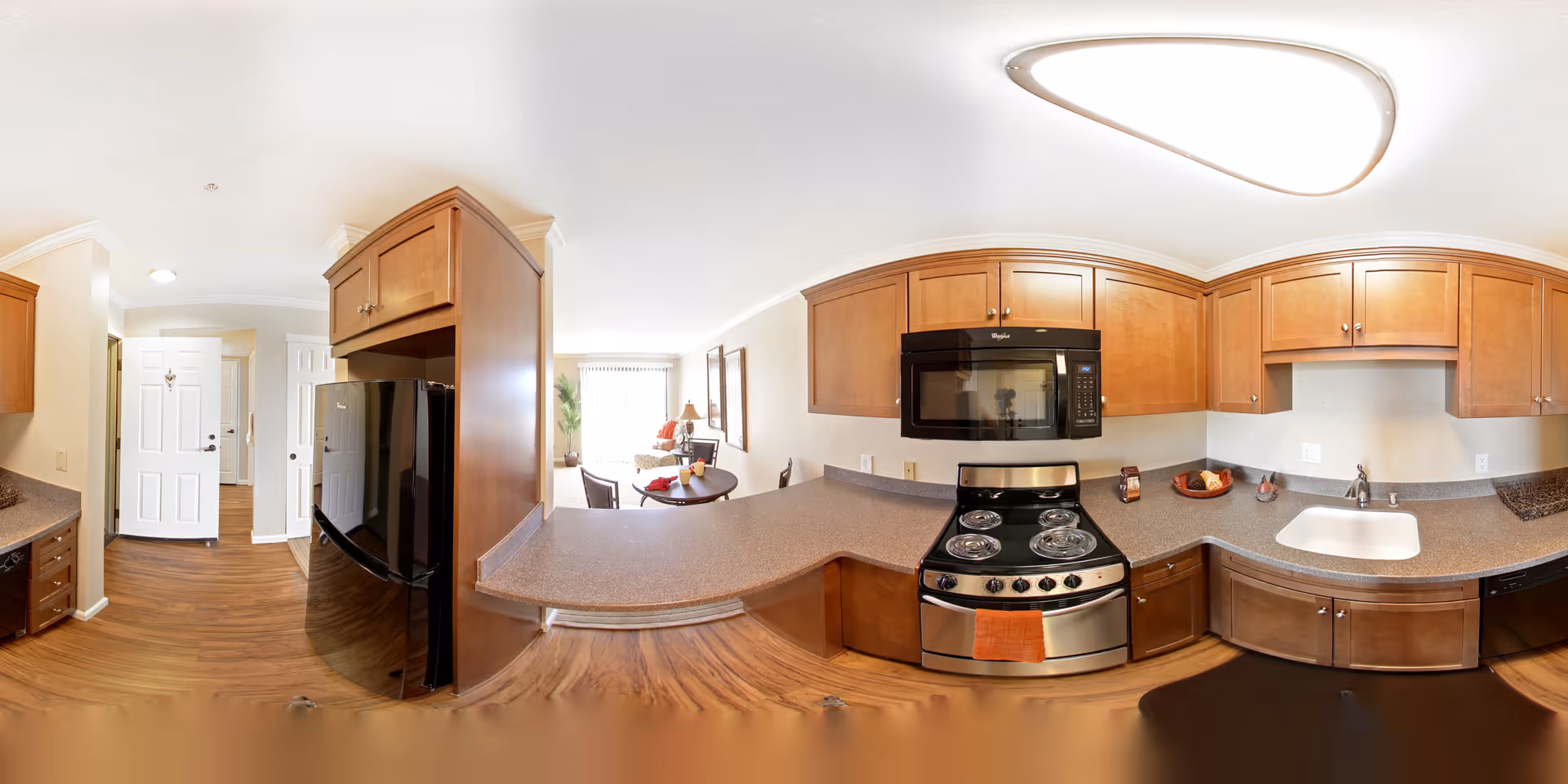 A bright and modern kitchen with wooden cabinets, a black refrigerator, a stainless steel stove with an orange towel hanging on the handle, a microwave above the stove, a white sink, and a countertop extending into a small dining area with a round table and chairs. The kitchen opens into a living space with large windows letting in natural light.