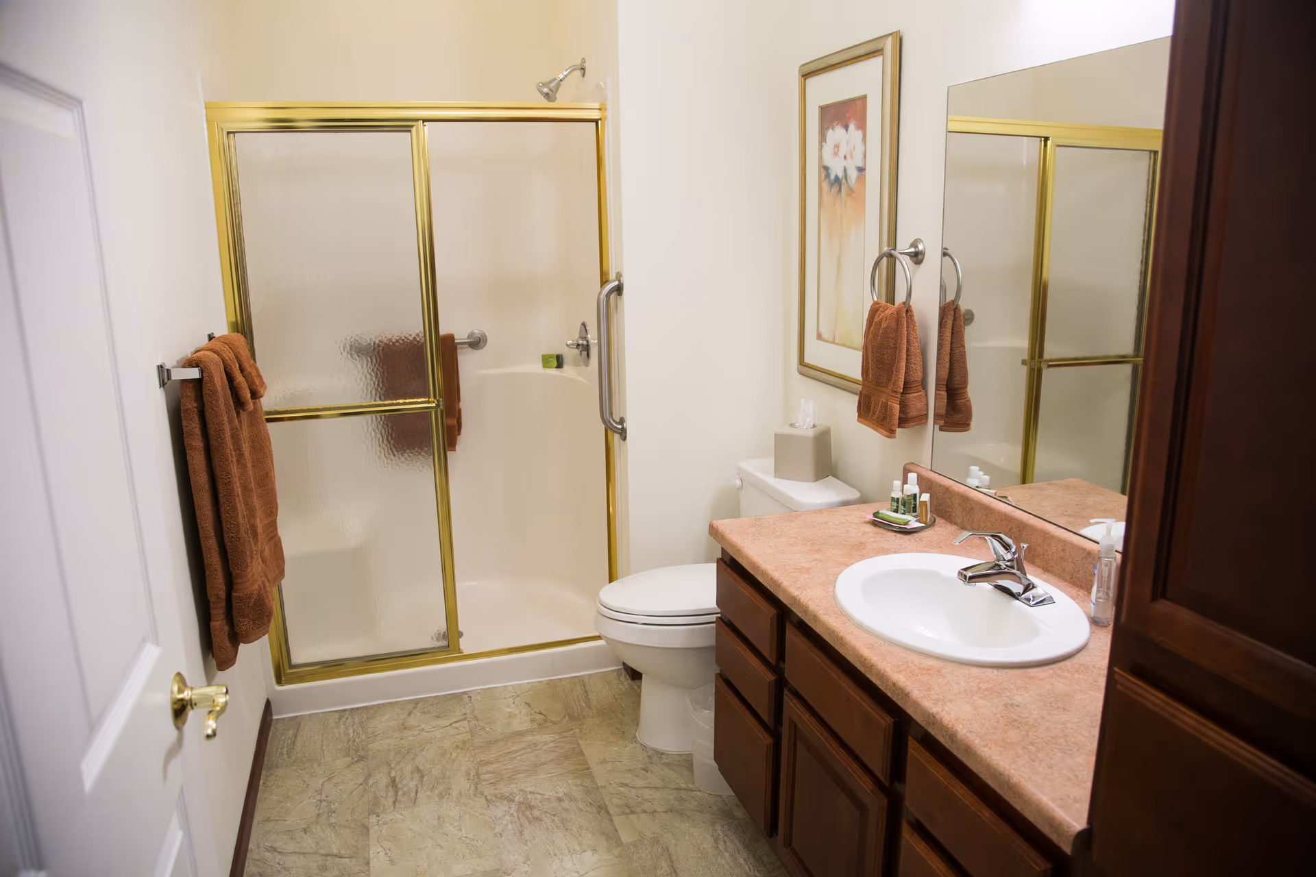 A bathroom with a shower enclosed by frosted glass doors with gold trim, a toilet, a sink with a pinkish countertop, wooden cabinets, a large mirror, brown towels hanging on a towel rack and ring, and a framed floral picture on the wall.