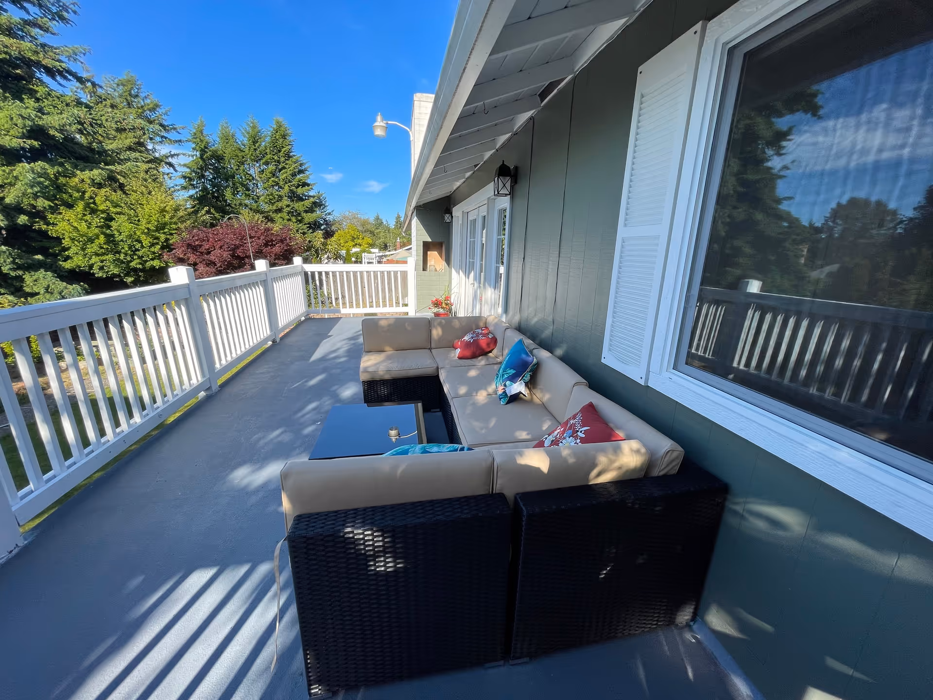 Outdoor balcony area with a beige cushioned sectional sofa and colorful pillows, a small black coffee table, white railing, and green trees in the background under a clear blue sky.