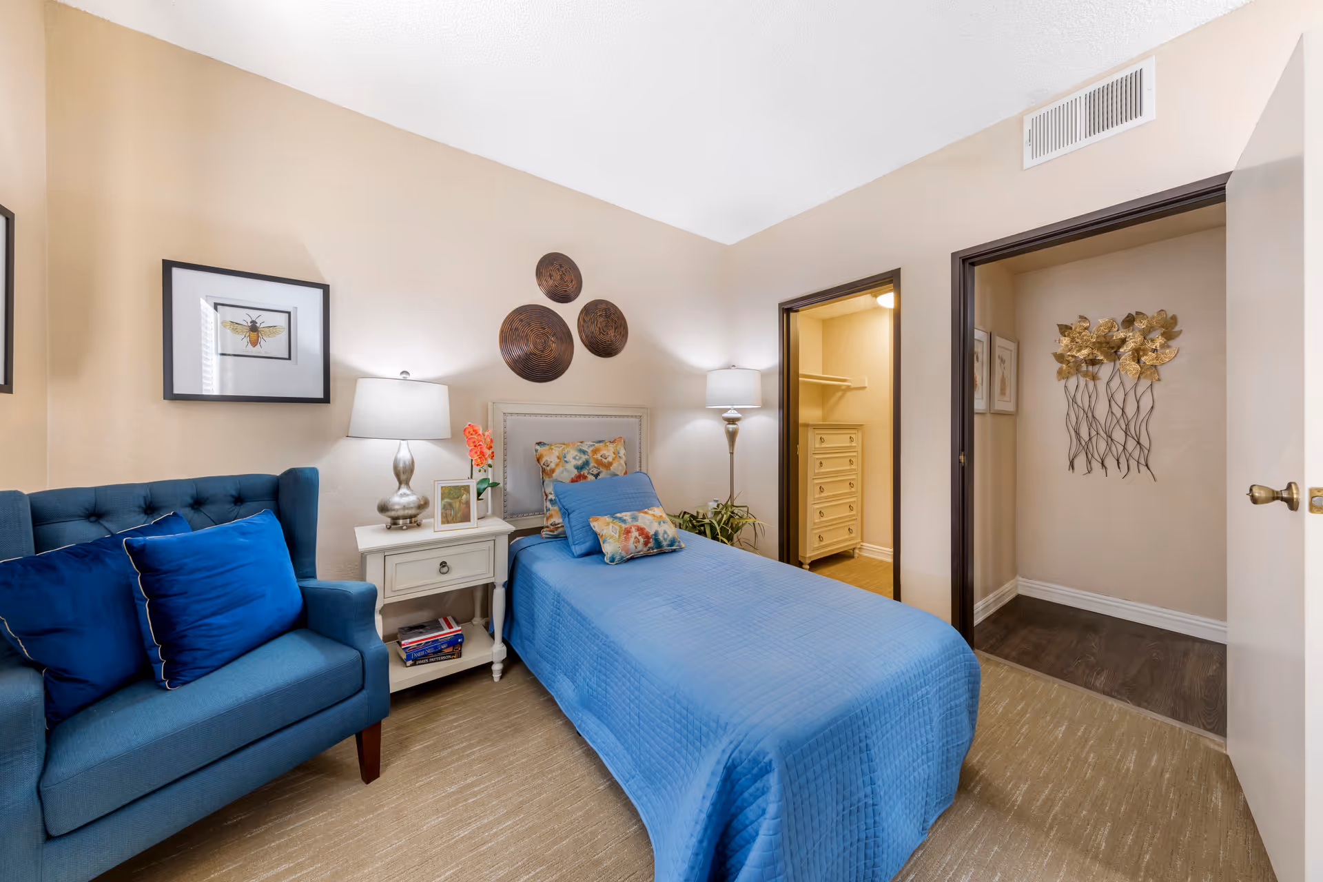 A cozy bedroom with a single bed covered in a blue quilt and floral pillows. Next to the bed is a white nightstand with a lamp, a small plant, and framed pictures. A blue upholstered armchair with two blue pillows is positioned to the left. The walls are beige with decorative wall art, including three circular wooden pieces above the bed and a framed insect illustration. There is an open doorway leading to a walk-in closet with a dresser inside, and another open doorway showing a wall with metallic floral wall decor.