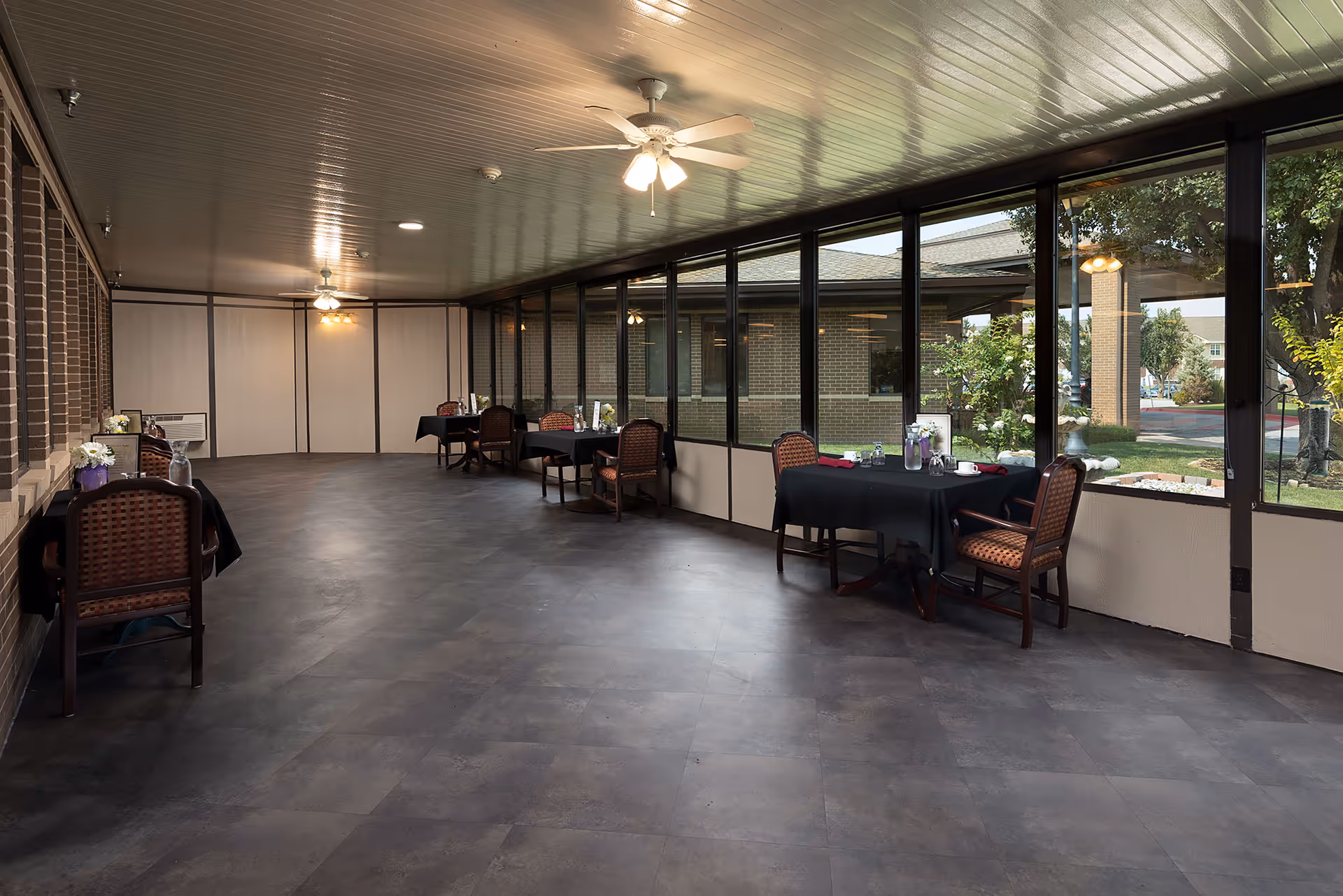 Enclosed sunroom-style common room with several small dining tables and chairs along large windows overlooking the facility courtyard.