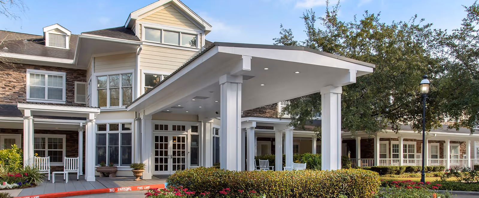 Covered entrance and porte-cochere of a senior living facility with white columns, rocking chairs, and landscaped shrubs.