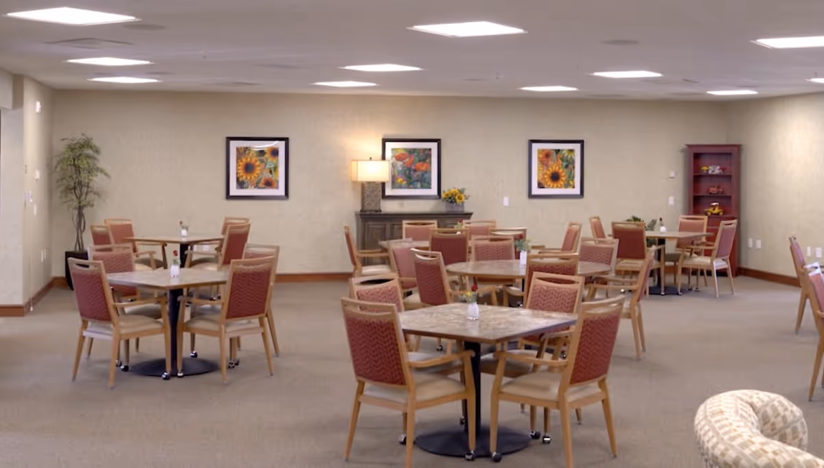 A spacious dining room with multiple square and round tables, each surrounded by four wooden chairs with red cushioned seats. The walls are beige with three framed floral paintings and a lamp on a sideboard. There is a tall plant in the corner and a wooden shelving unit with decorative items. The room is well-lit with ceiling lights.