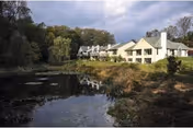 Multi-unit senior living building beside a pond with reflections and wooded surroundings under a cloudy sky.