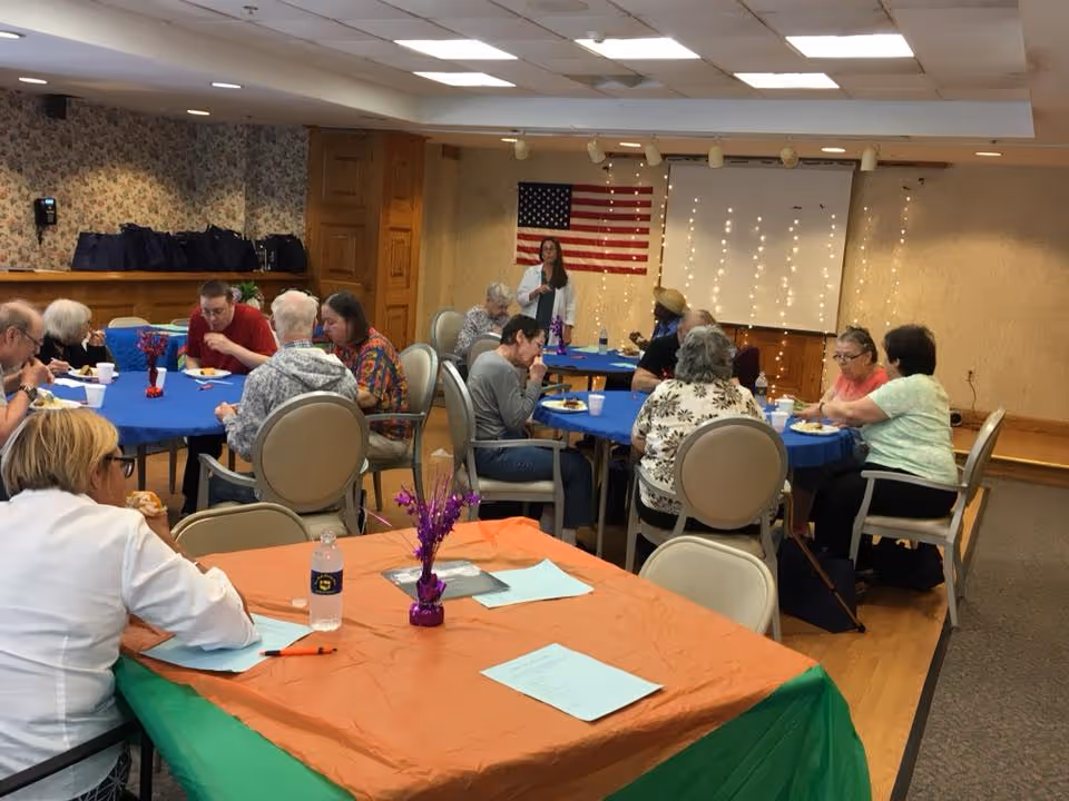 Seniors seated at decorated tables in a community dining/activity room with an American flag and a woman speaking at the front.