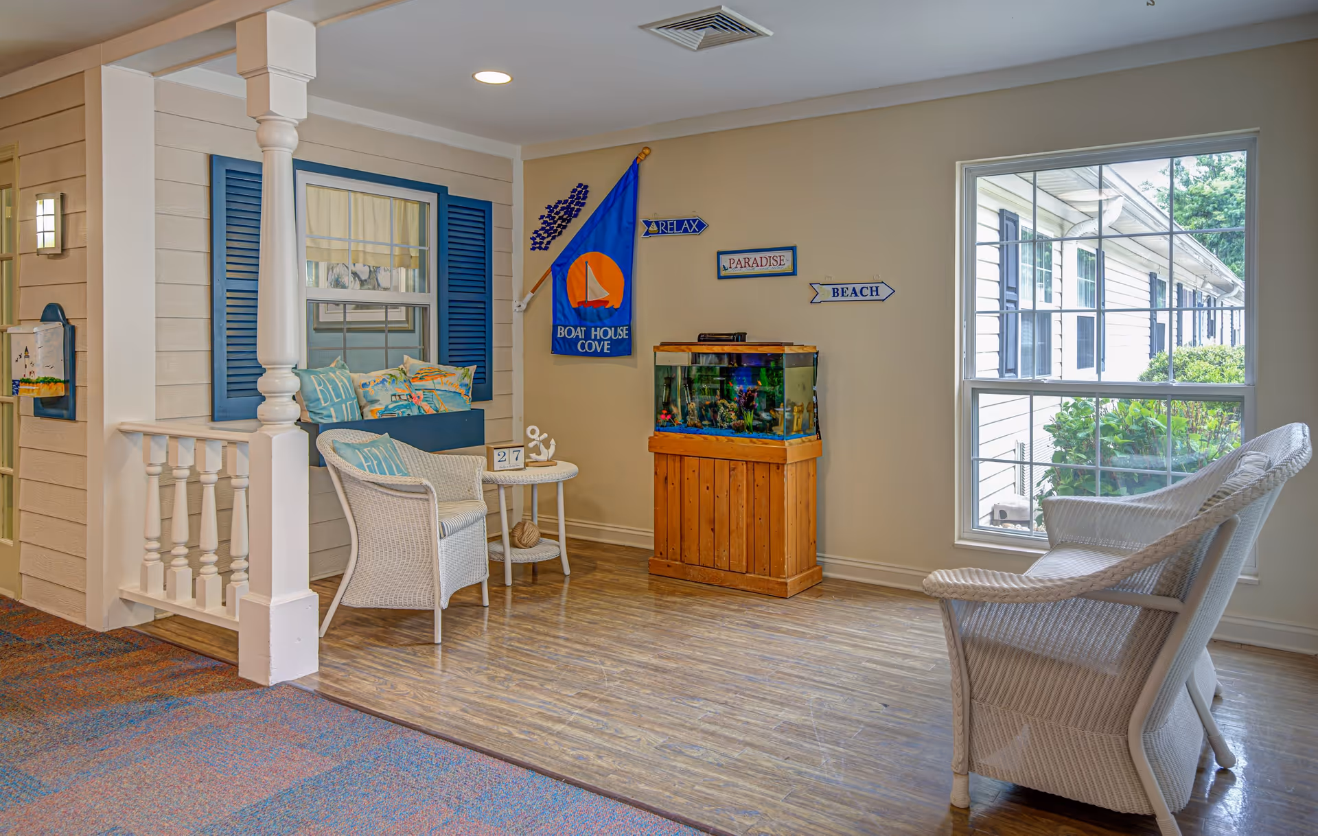 A cozy indoor sitting area with a nautical theme, featuring white wicker chairs and a small round table with decorative items. There is a wooden fish tank stand with a fish tank on top, and wall decorations including a blue flag with a sailboat and signs that say 'RELAX', 'PARADISE', and 'BEACH'. A large window lets in natural light and shows greenery outside. The floor is a combination of wood and carpet, and the walls are painted beige.