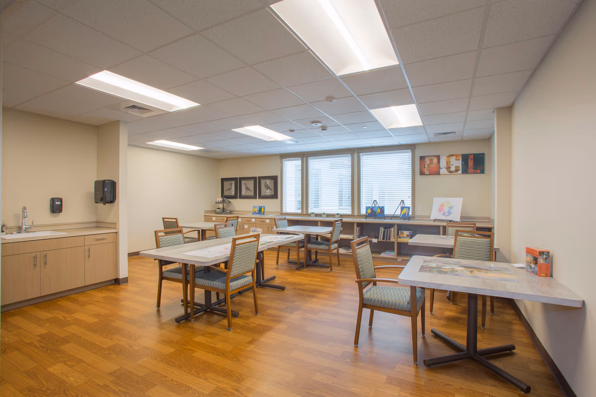 Well-lit communal activity/dining room with several tables and chairs, cabinets and a sink along one wall, and windows with blinds.