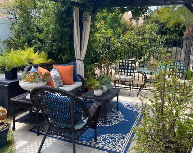 Outdoor patio area with wicker furniture including a loveseat and chair with cushions and colorful throw pillows, a coffee table with potted plants, a blue patterned outdoor rug, and a metal table with two chairs with striped cushions. The area is surrounded by lush greenery and plants, with a white curtain hanging from a pergola.