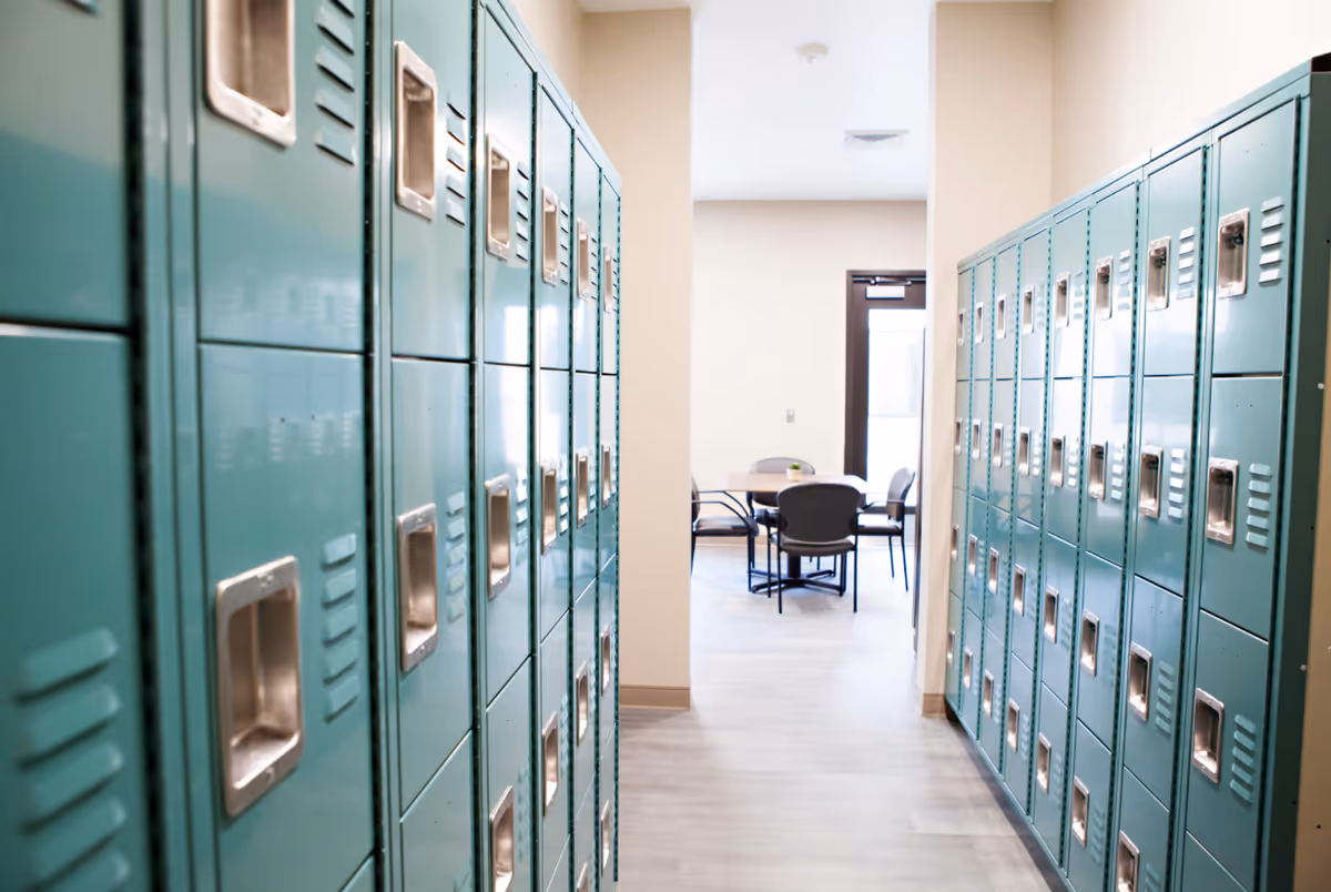A hallway lined with teal lockers on both sides leading to a room with a round table and four chairs near a door with a window.