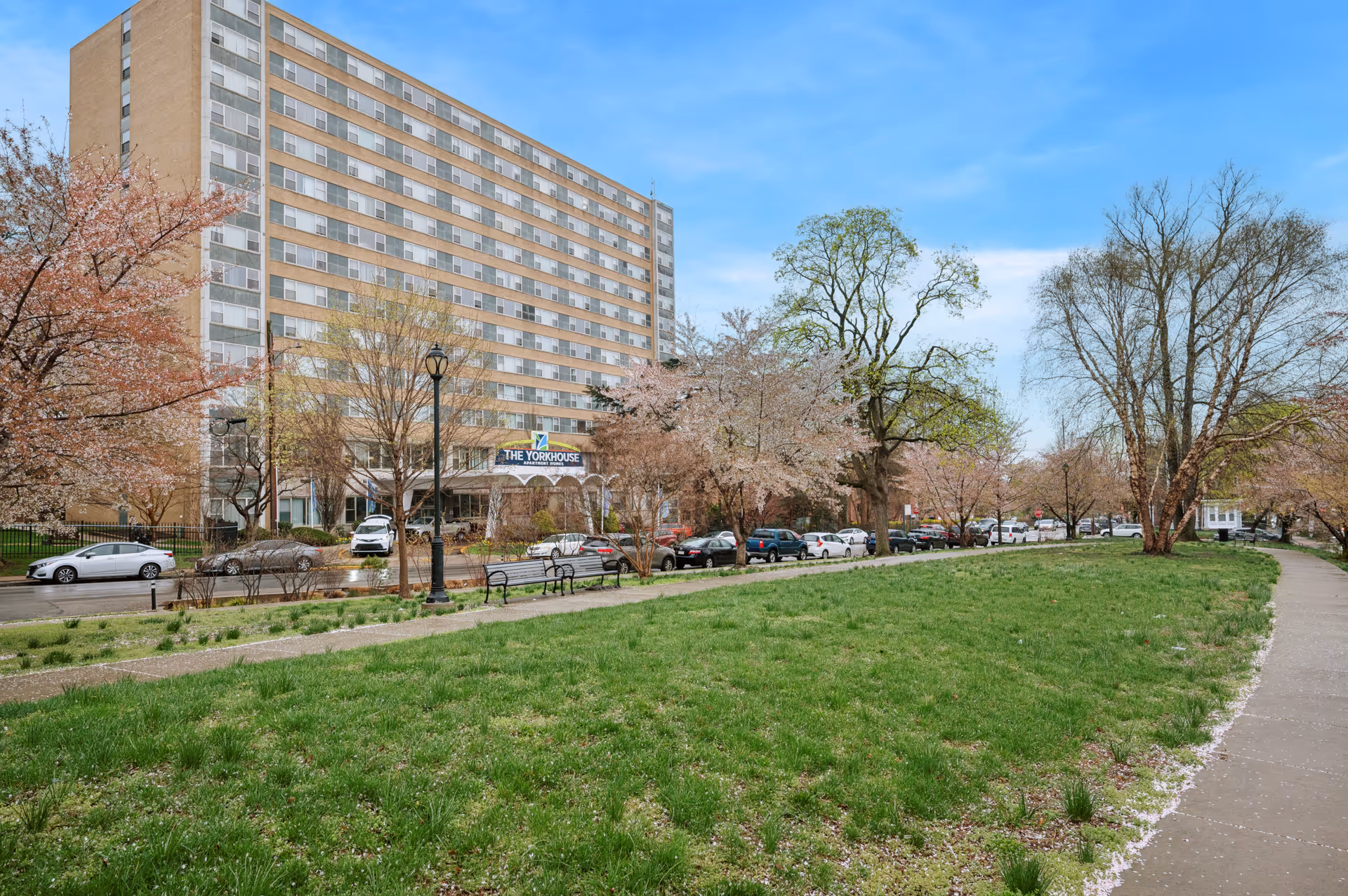 A multi-story residential building named The Yorkhouse with a green lawn and sidewalk in the foreground, surrounded by trees and parked cars along the street under a blue sky.