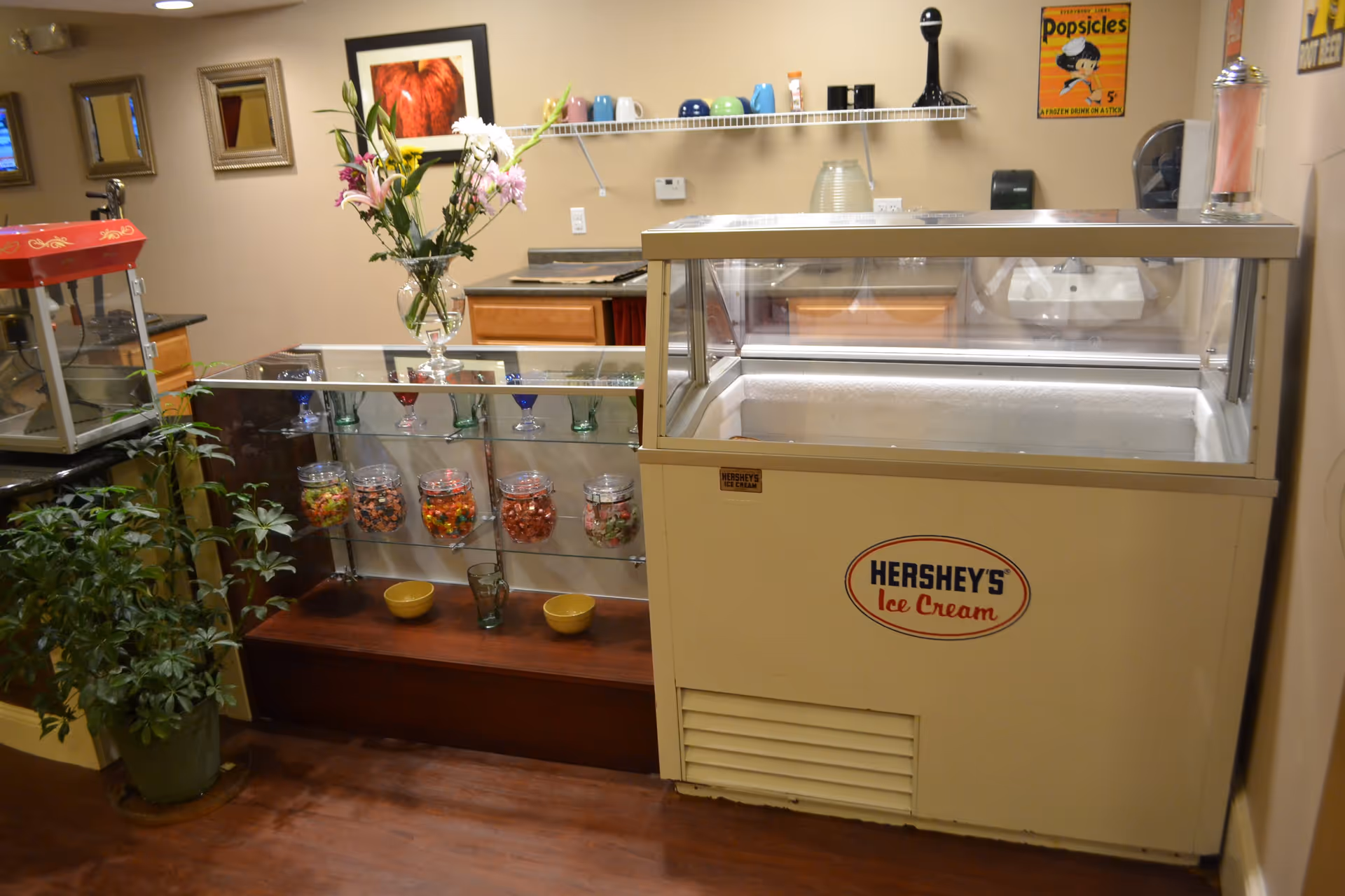 Interior view of a snack and dessert area featuring a Hershey's Ice Cream freezer, a glass display case with jars of candy, a popcorn machine, a vase with flowers, and a shelf with colorful mugs on a beige wall.
