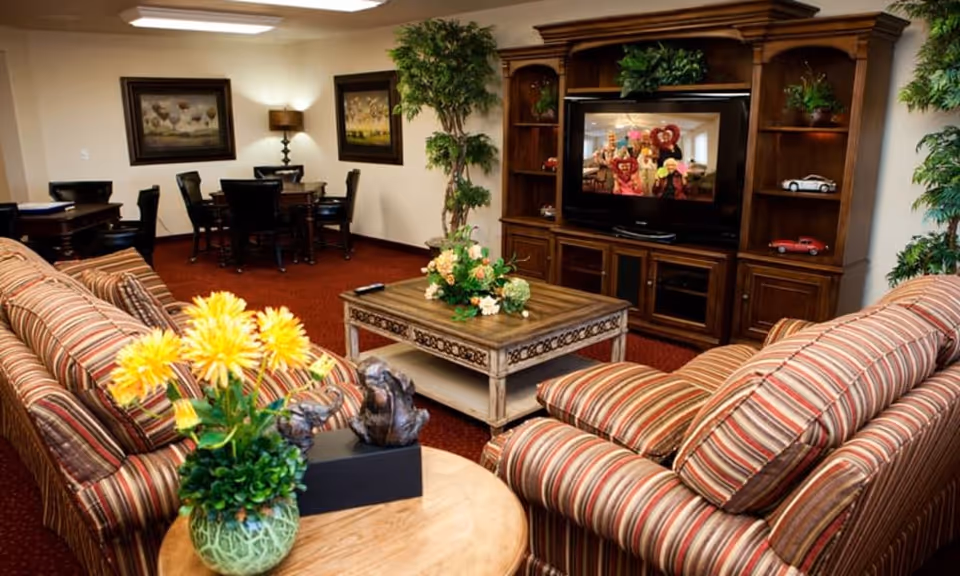 A cozy living room area with striped sofas facing a wooden entertainment center with a TV. The TV screen shows a group of people holding heart-shaped decorations. There is a wooden coffee table with a floral arrangement in the center, and a round side table with a yellow flower arrangement in the foreground. In the background, there are two framed paintings on the wall, a lamp, and a dining table with chairs. The room has warm lighting and a red carpeted floor.