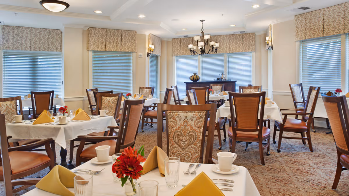 Bright dining room with multiple set tables, wooden chairs, folded napkins and floral centerpieces.