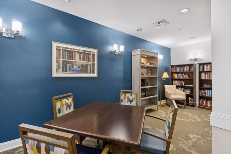 A cozy reading and meeting area with a wooden table surrounded by four chairs with patterned cushions. The walls are painted blue and white, adorned with framed artwork of bookshelves. There are multiple bookshelves filled with books, a beige armchair, and a floor lamp providing warm lighting. The carpet has a subtle floral pattern.