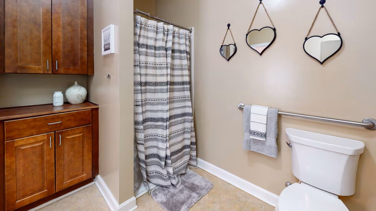 Bathroom featuring wooden cabinets, a shower with a patterned curtain, a toilet with a grab bar and towel, and three heart-shaped mirrors on a beige wall.