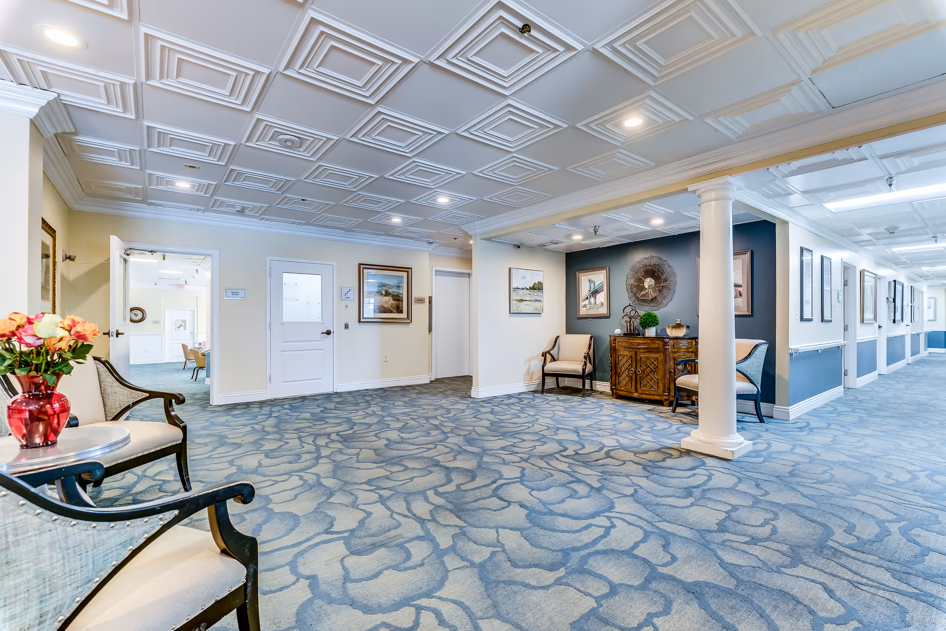 A spacious, well-lit hallway in a senior living facility with patterned blue carpet, white paneled ceiling, and cream-colored walls. The hallway features decorative columns, framed artwork, and seating areas with chairs and a small table holding a red vase with flowers. Doors and a corridor are visible in the background.