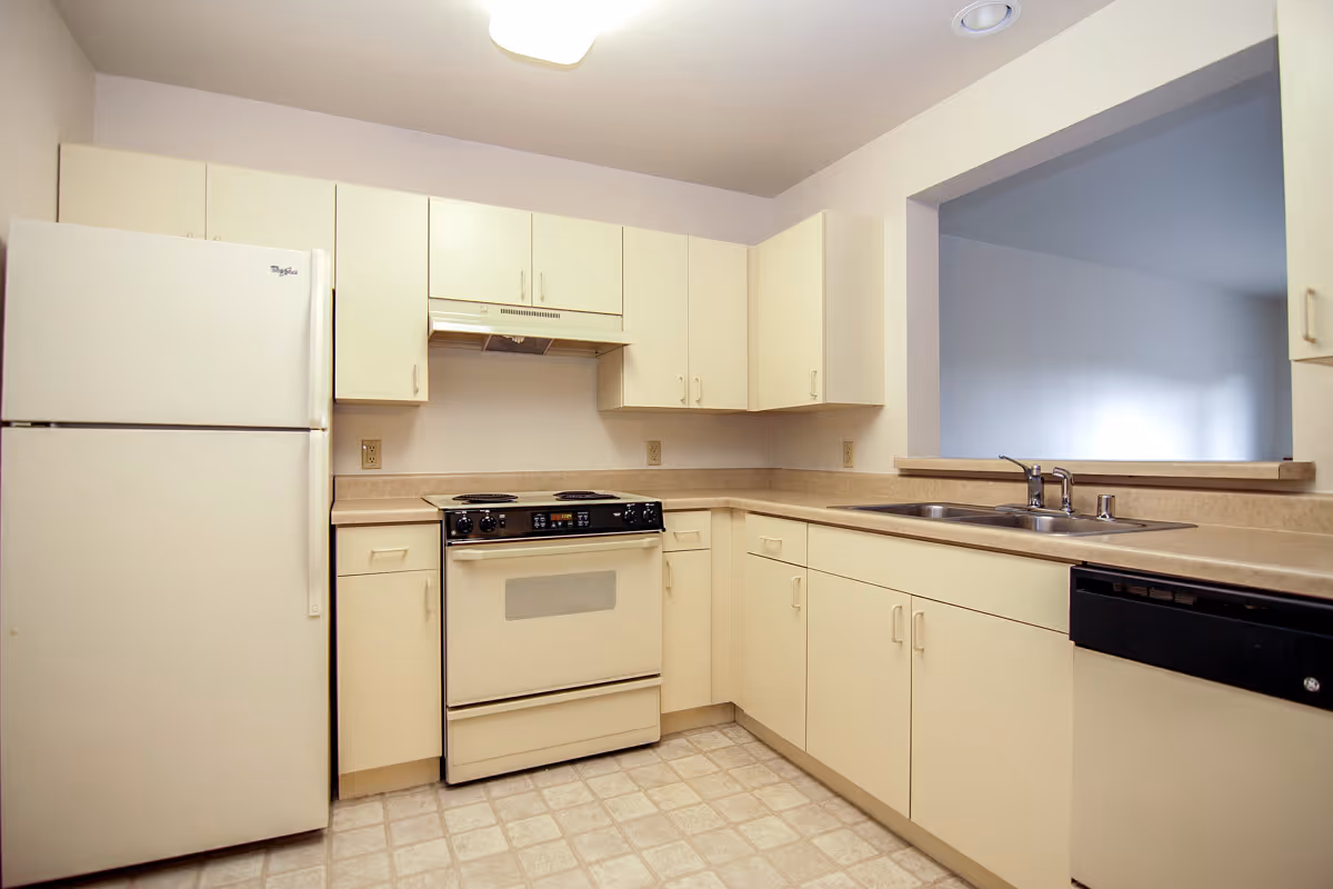 A clean kitchen with beige cabinets and countertops, featuring a white refrigerator, an electric stove with oven, a double sink, and a dishwasher. The kitchen has a tiled floor and a pass-through window opening to another room.