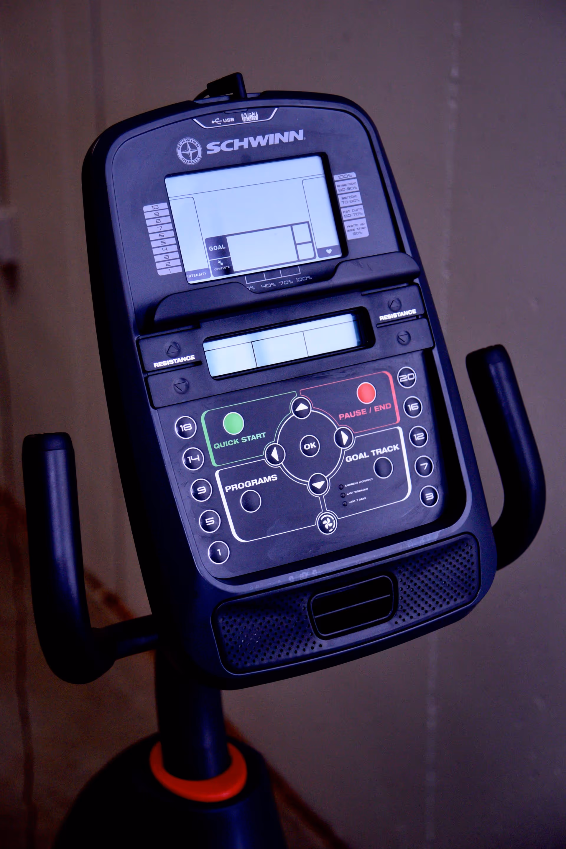 Close-up view of the control panel of a Schwinn exercise bike with buttons for quick start, pause/end, programs, and goal tracking, along with resistance adjustment knobs.