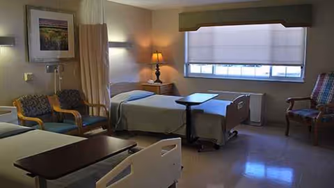 A hospital-style patient room with two beds, chairs, and a window view.