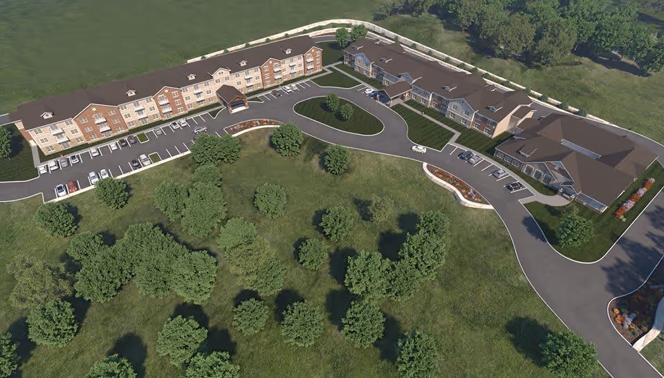 Aerial view of The Castlewood Senior Living facility showing a large, U-shaped building surrounded by green lawns and trees. The building has multiple parking spaces and a driveway leading to the entrance.