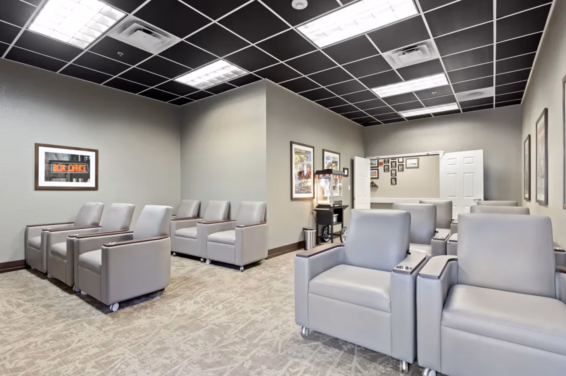 A room with multiple rows of gray cushioned armchairs on wheels arranged facing forward, resembling a small theater or screening room. The walls are light gray with framed posters and a popcorn machine near the back. The ceiling has a black grid pattern with fluorescent lighting.