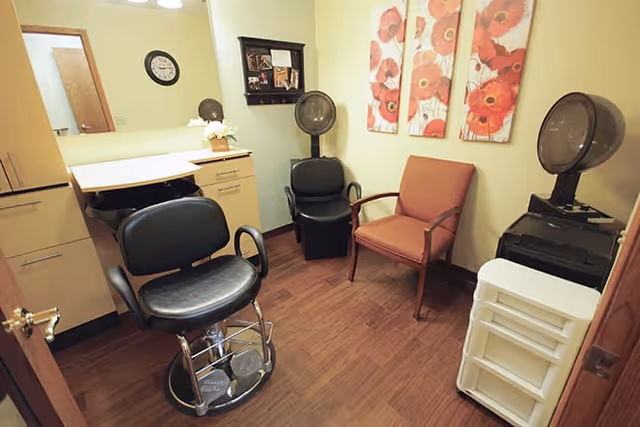 Small room with salon-style chairs, two hair dryers mounted on stands, a cabinet with drawers, a wall clock, and three floral paintings on the wall.
