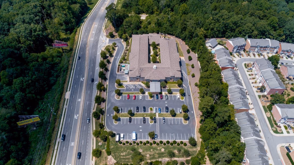 Aerial view of the Ivy Springs at Buford senior living building with its parking lot beside a multi-lane road and neighboring homes and trees.