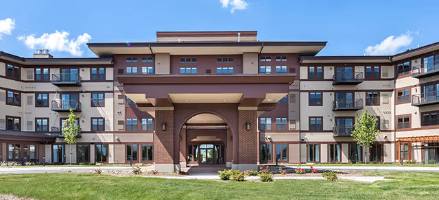 Front exterior view of a modern senior living facility building with multiple windows, balconies, and a covered entrance. The building is surrounded by a well-maintained lawn and small trees under a blue sky with some clouds.