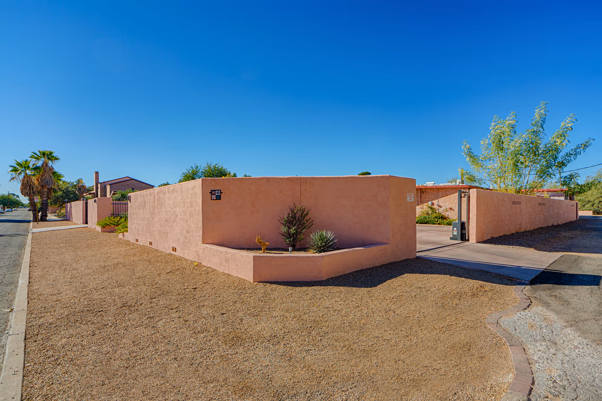 Low pink stucco wall and gated driveway leading into a desert-style residential property with gravel landscaping and palm trees under a clear blue sky.