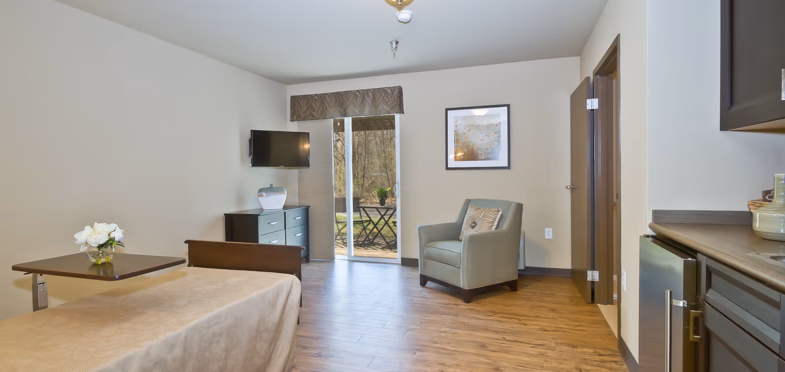 Sunlit studio room with a bed, armchair, wall-mounted TV on a dresser, kitchenette at the right, and a sliding glass door to an outdoor patio.