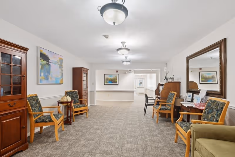 A well-lit hallway in a senior living facility with carpeted floor, wooden chairs with patterned cushions, wooden cabinets, a small table with a globe, framed artwork on the walls, a large mirror, and ceiling light fixtures.
