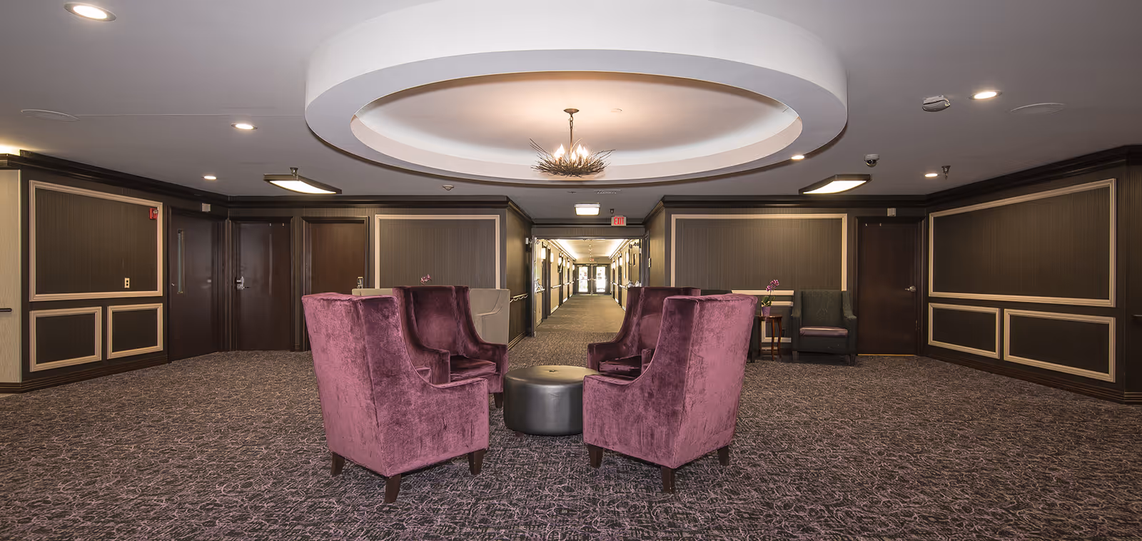 A spacious interior common area with four purple upholstered armchairs arranged around a small round black ottoman. The room features patterned carpet, dark wood paneling with beige trim, and a circular recessed ceiling with a modern chandelier. A hallway with doors and an exit sign is visible in the background.