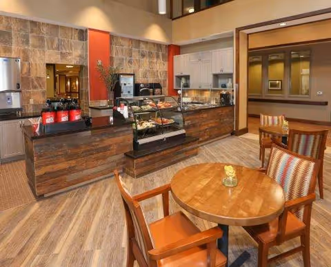 Interior view of a dining area in a nursing home featuring a wooden round table with four chairs, a counter with a glass display case containing food items, coffee dispensers, and a kitchen area in the background with cabinets and appliances.