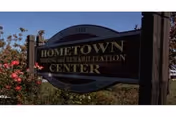 Decorative wooden entrance sign reading 'Hometown Nursing and Rehabilitation Center' beside flowering shrubs under a clear blue sky.