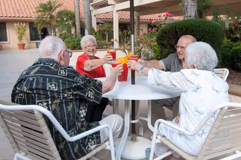 Four elderly people sitting around a white outdoor table, raising their glasses in a toast. They are smiling and enjoying drinks with orange slices as garnish. The setting is a sunny outdoor patio with plants and a building in the background.