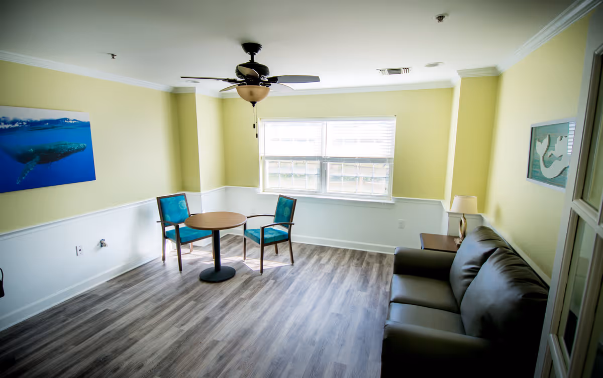 A bright living room with light yellow walls and wood flooring. The room features a ceiling fan with light, a window with blinds, a small round table with two blue cushioned chairs, a dark brown leather sofa, a side table with a lamp, and two wall decorations depicting whales.